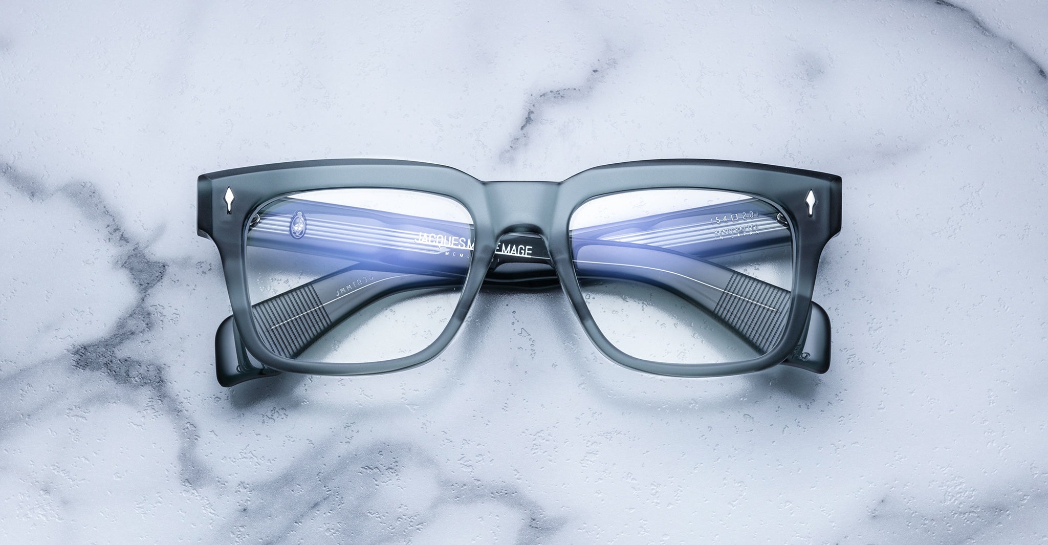 The Jacques Marie Mage Torino rectangular eyeglass frames, handcrafted from acetate by Jacques Marie Mage, rest on a white marble surface with reflections visible on their lenses.
