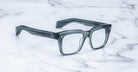Jacques Marie Mage Torino eyewear: clear gray handcrafted acetate frames with bold rectangular design, inspired by the Jacques Marie Mage aesthetic, shown on a white marble surface.