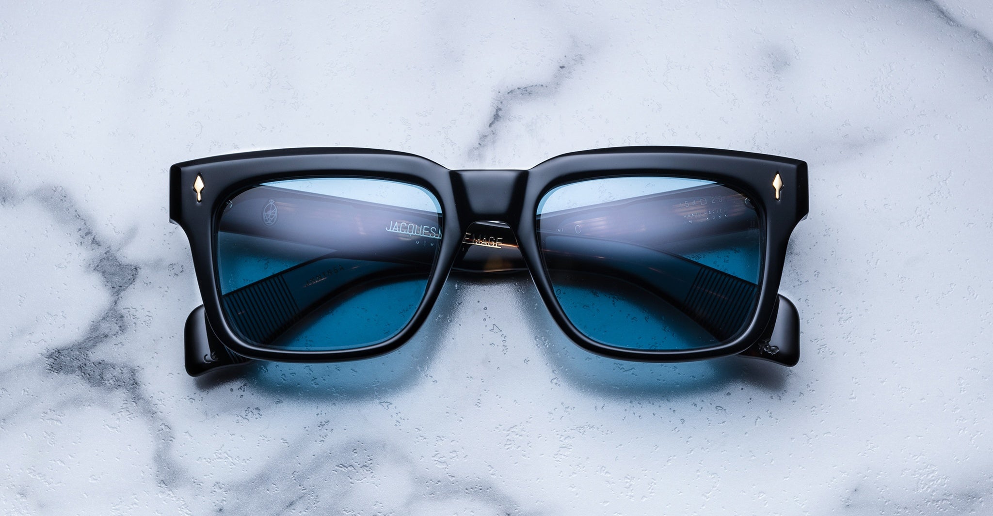 A pair of Jacques Marie Mage Torino sunglasses by Jacques Marie Mage, featuring black frames and blue-tinted lenses, rests on a white marble surface.