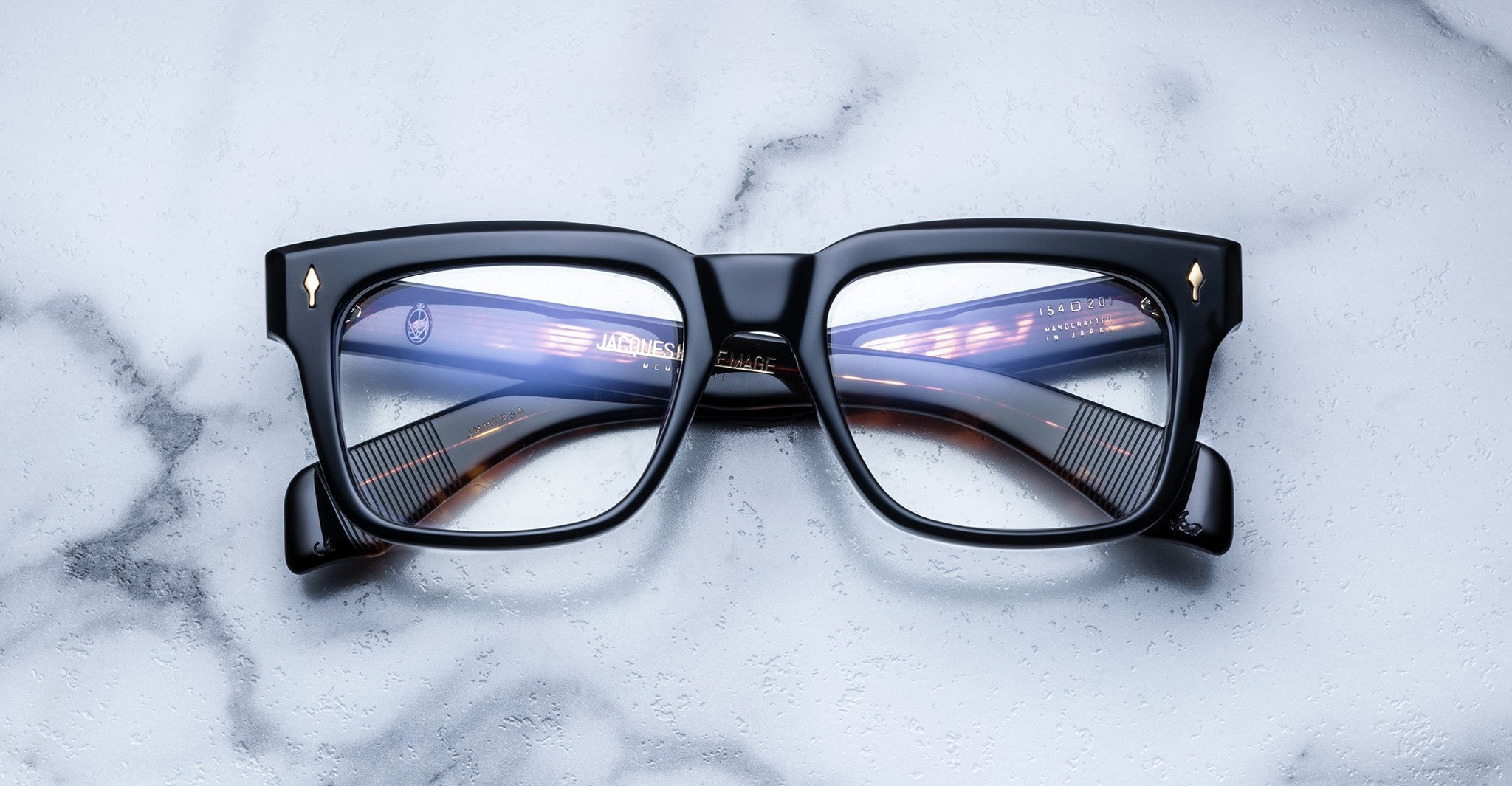 Jacques Marie Mage Torino eyeglasses feature black bold rectangular frames with gold corner accents, styled by Jacques Marie Mage, displayed on a white marble surface.