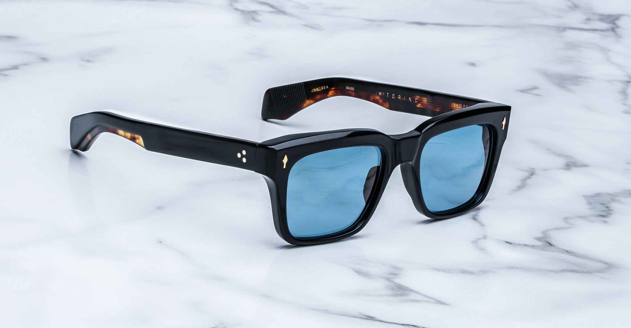 The Jacques Marie Mage Torino sunglasses by Jacques Marie Mage feature bold black square frames and blue tinted lenses, elegantly displayed on a white marble surface.
