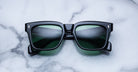 The Jacques Marie Mage Torino by Jacques Marie Mage features black bold rectangular acetate frames with green-tinted lenses, displayed on white marble and highlighting sophisticated handcrafted eyewear.