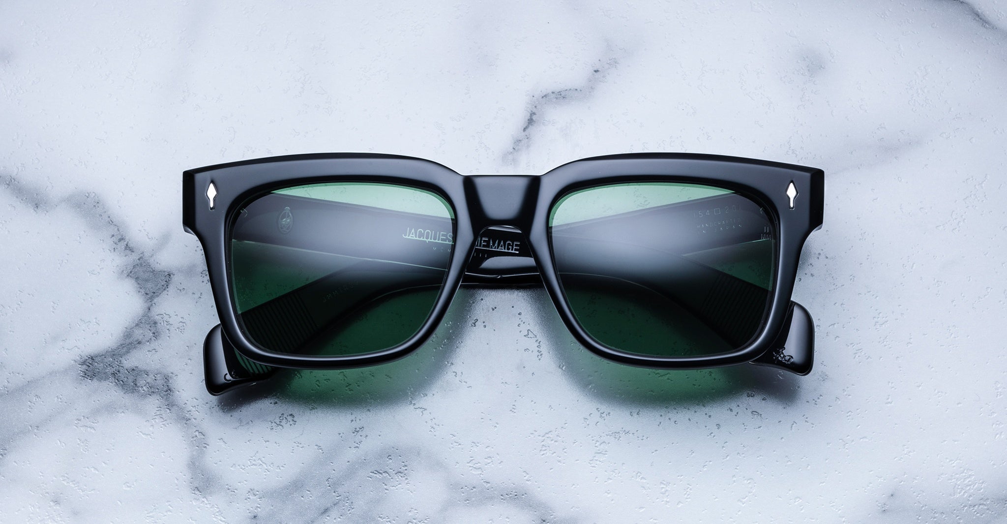 The Jacques Marie Mage Torino by Jacques Marie Mage features black bold rectangular acetate frames with green-tinted lenses, displayed on white marble and highlighting sophisticated handcrafted eyewear.