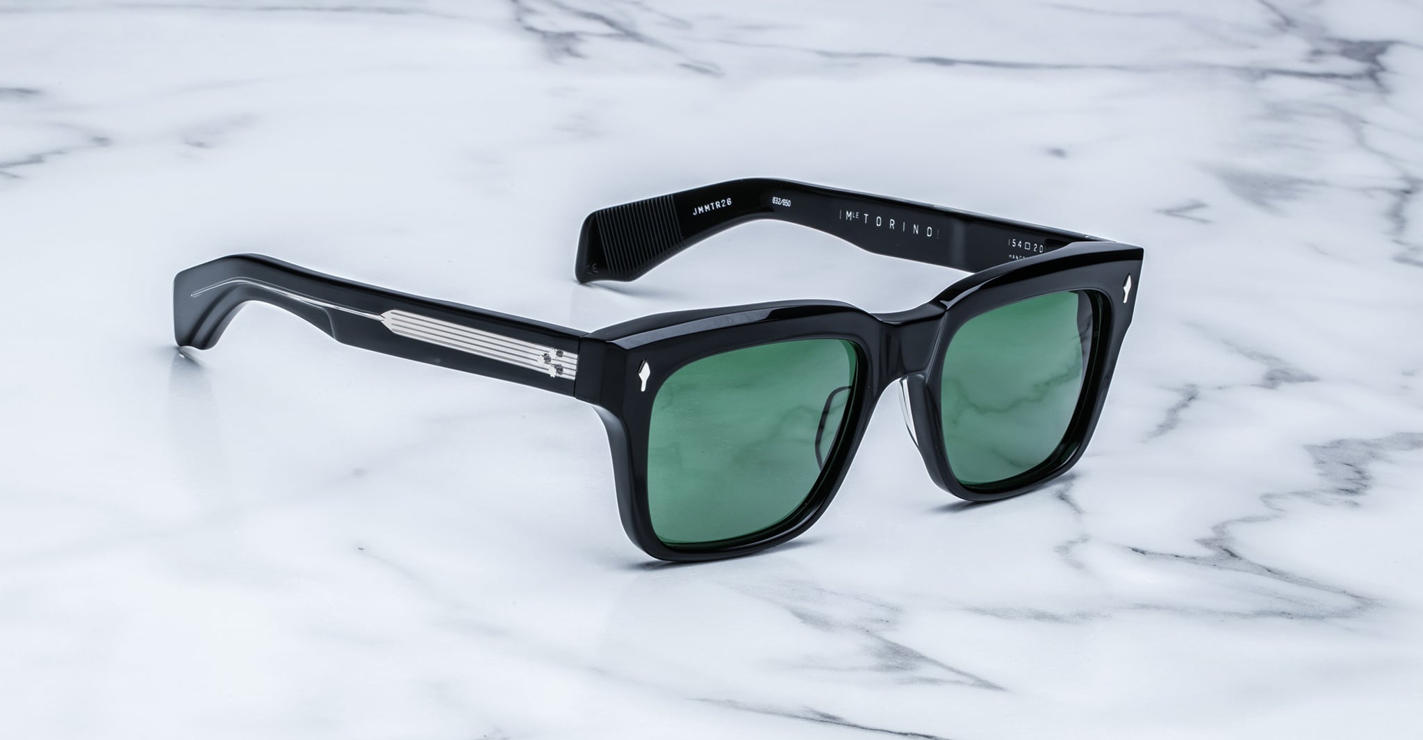 The Jacques Marie Mage Torino sunglasses by Jacques Marie Mage feature bold black rectangular acetate frames with green lenses, elegantly displayed on a white marble surface.