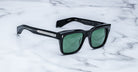 The Jacques Marie Mage Torino sunglasses by Jacques Marie Mage feature bold black rectangular acetate frames with green lenses, elegantly displayed on a white marble surface.