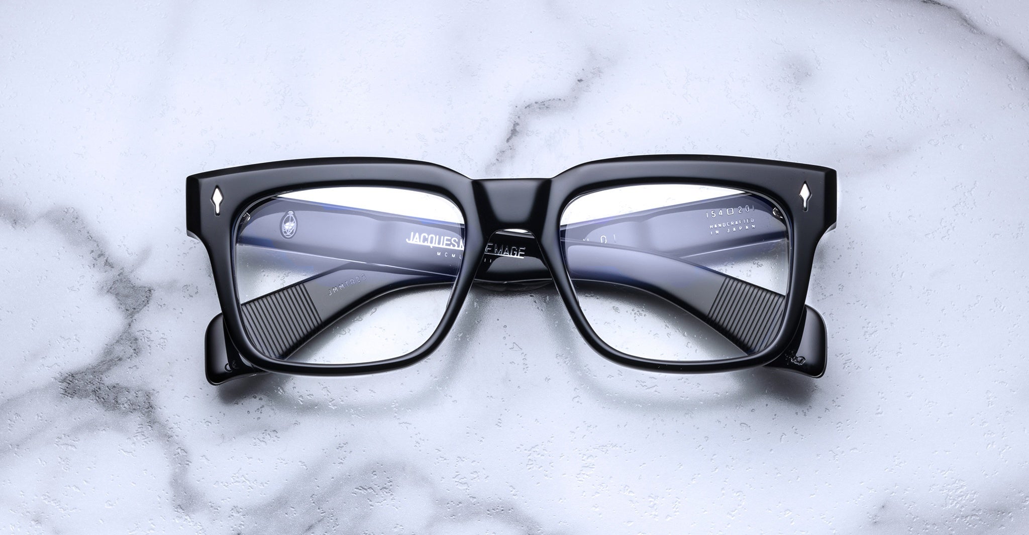 The Jacques Marie Mage Torino by Jacques Marie Mage features black rectangular frames made from handcrafted acetate, showcased on a white marble surface and viewed from above.