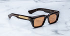 Black rectangular sunglasses with thick frames and brown lenses, featuring gold accents on the temples, displayed on a white marble surface.