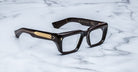 Thick, rectangular, Walker dark brown eyeglass frames with gold detailing on the temples rest on a white marble surface.