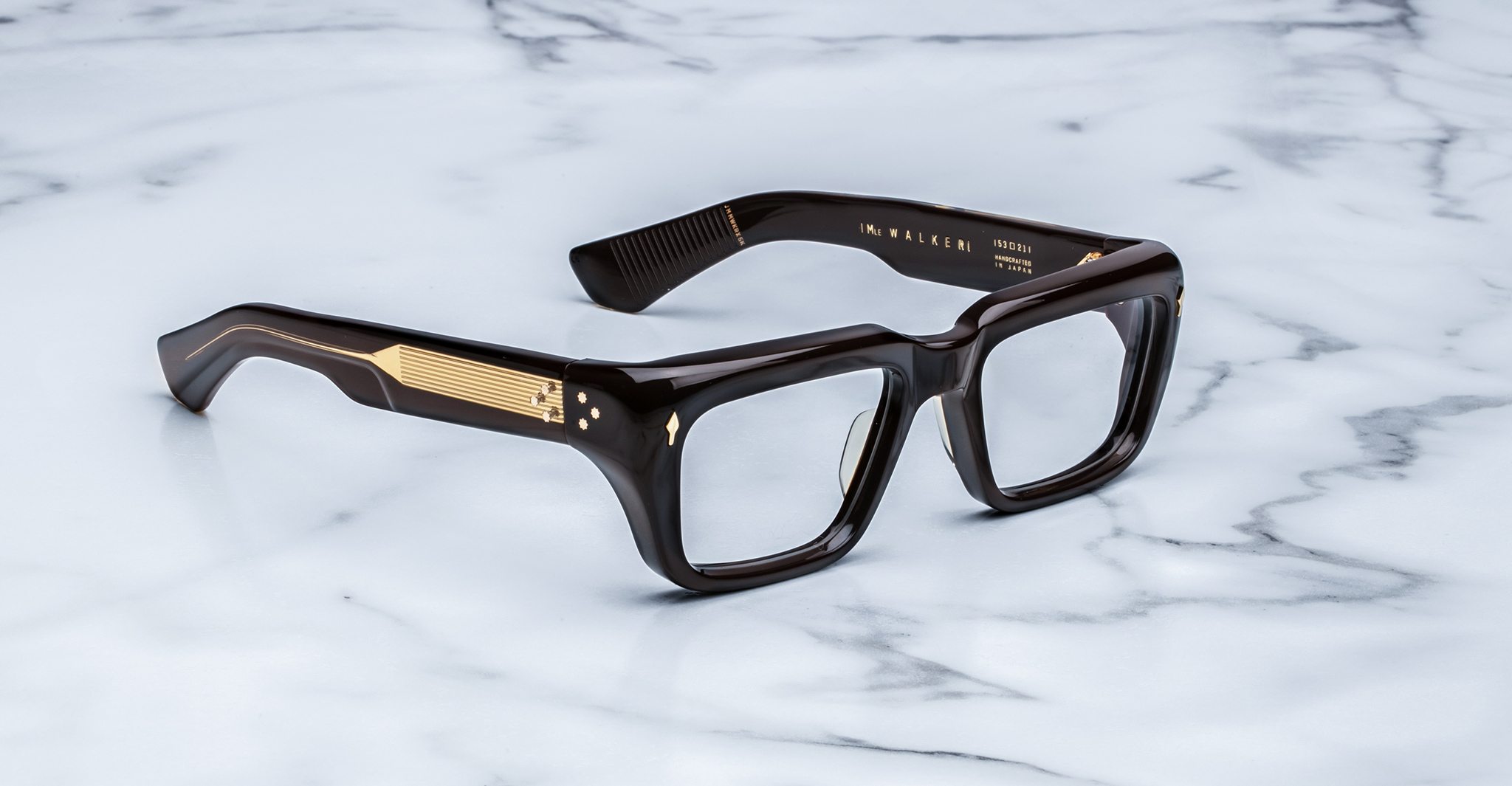 Thick, rectangular, Walker dark brown eyeglass frames with gold detailing on the temples rest on a white marble surface.