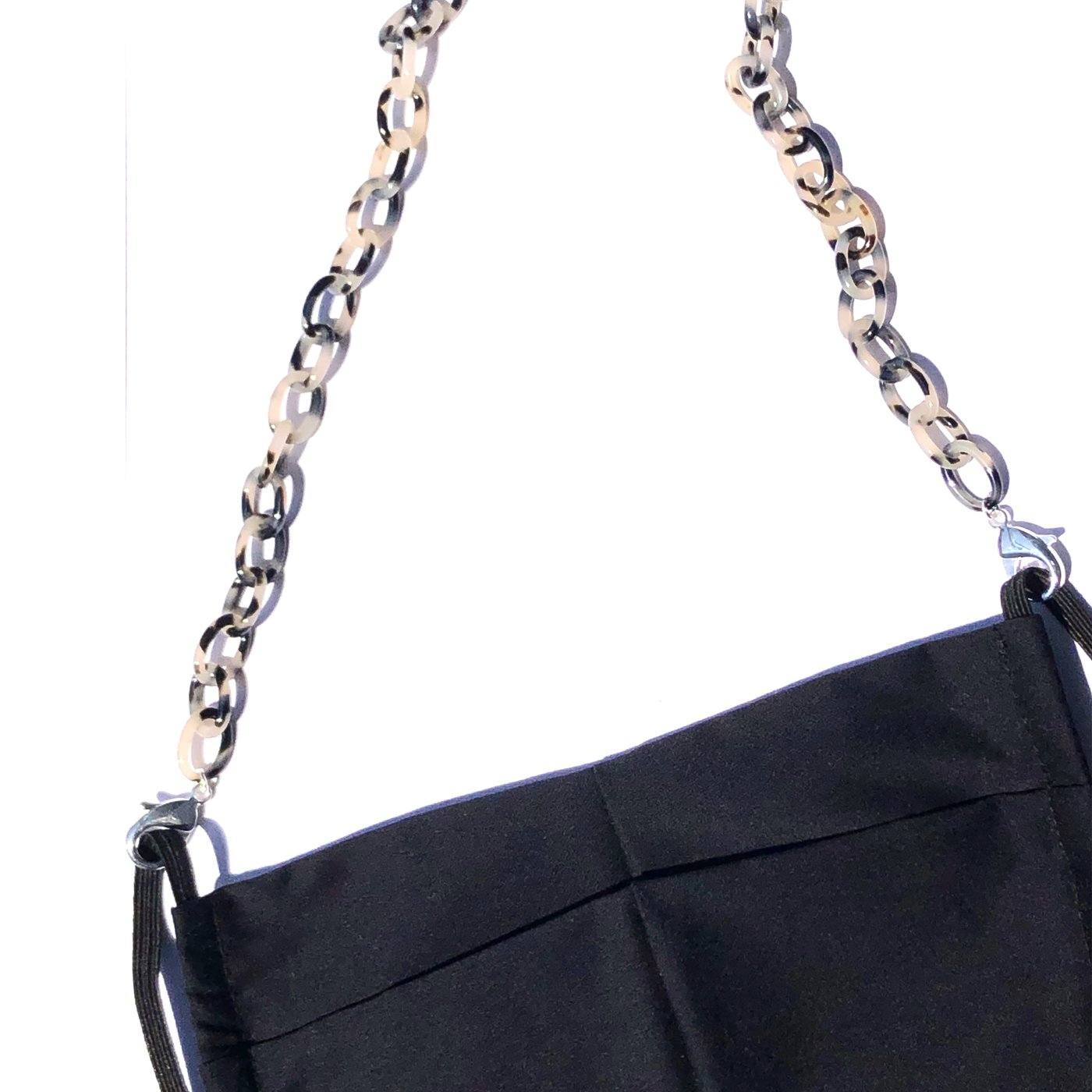 A close-up of a black cloth face mask with the Small Link Necklace Chain for Face Masks—a chunky, beige and black chain—attached to the ear loops, set against a white background.