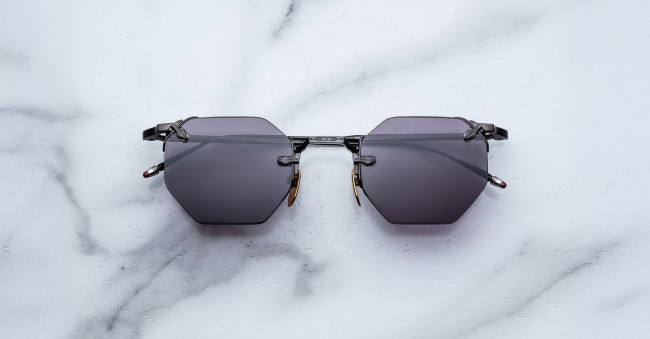 The Jacques Marie Mage El Dorado sunglasses, featuring geometric hexagonal black frames and dark lenses, are displayed on a white marble surface.