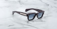 Jacques Marie Mage Fellini sunglasses with black square frames and silver temple detailing rest on a white marble surface. The lenses are dark-tinted, and branded info is engraved inside the arm.