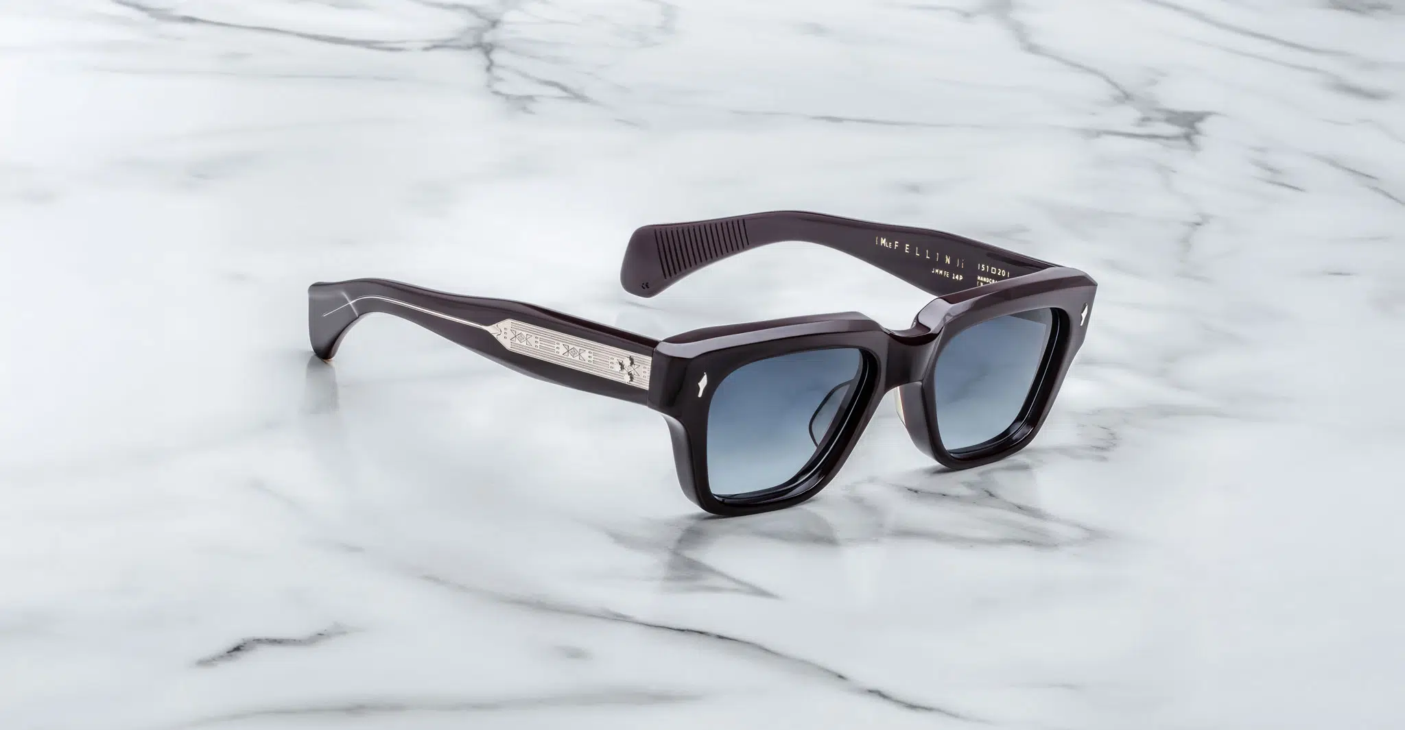 Jacques Marie Mage Fellini sunglasses with black square frames and silver temple detailing rest on a white marble surface. The lenses are dark-tinted, and branded info is engraved inside the arm.