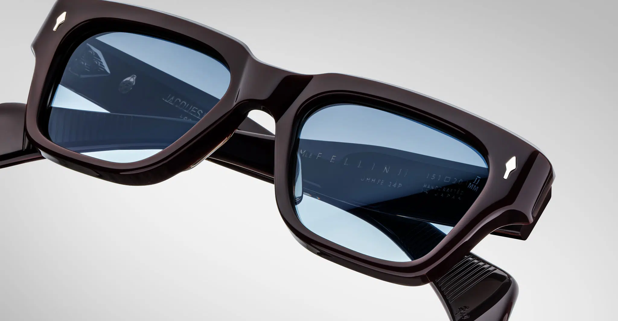 Close-up of Jacques Marie Mage Fellini eyeglasses with dark frames and light blue tinted lenses, highlighting the detailed frame and arms against a plain, light background.