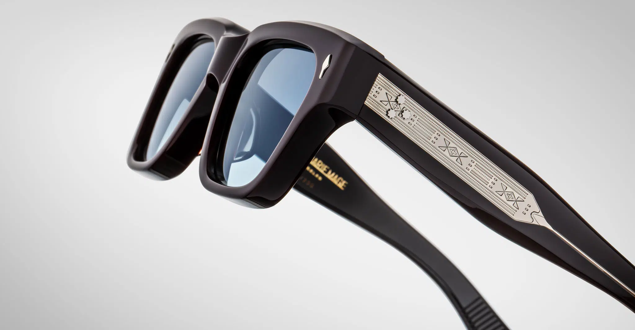 Close-up of Jacques Marie Mage Fellini black rectangular sunglasses with metallic inlays engraved with geometric patterns on the temples and blue-tinted lenses, shown against a plain background.