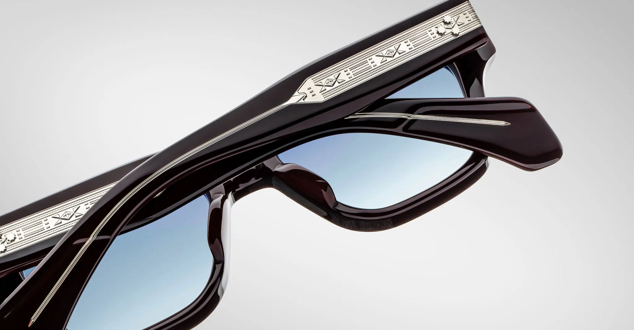 A close-up of folded Jacques Marie Mage Fellini sunglasses with dark frames, light blue tinted lenses, and silver temple detailing, set against a plain gray background.