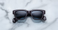 A pair of Jacques Marie Mage Fellini black rectangular sunglasses with dark lenses is laid flat on a white marble surface with gray veining.