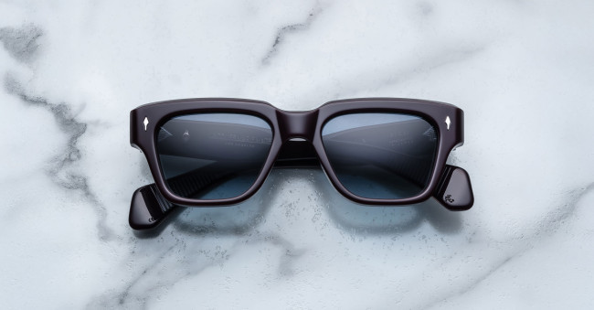 A pair of Jacques Marie Mage Fellini black rectangular sunglasses with dark lenses is laid flat on a white marble surface with gray veining.