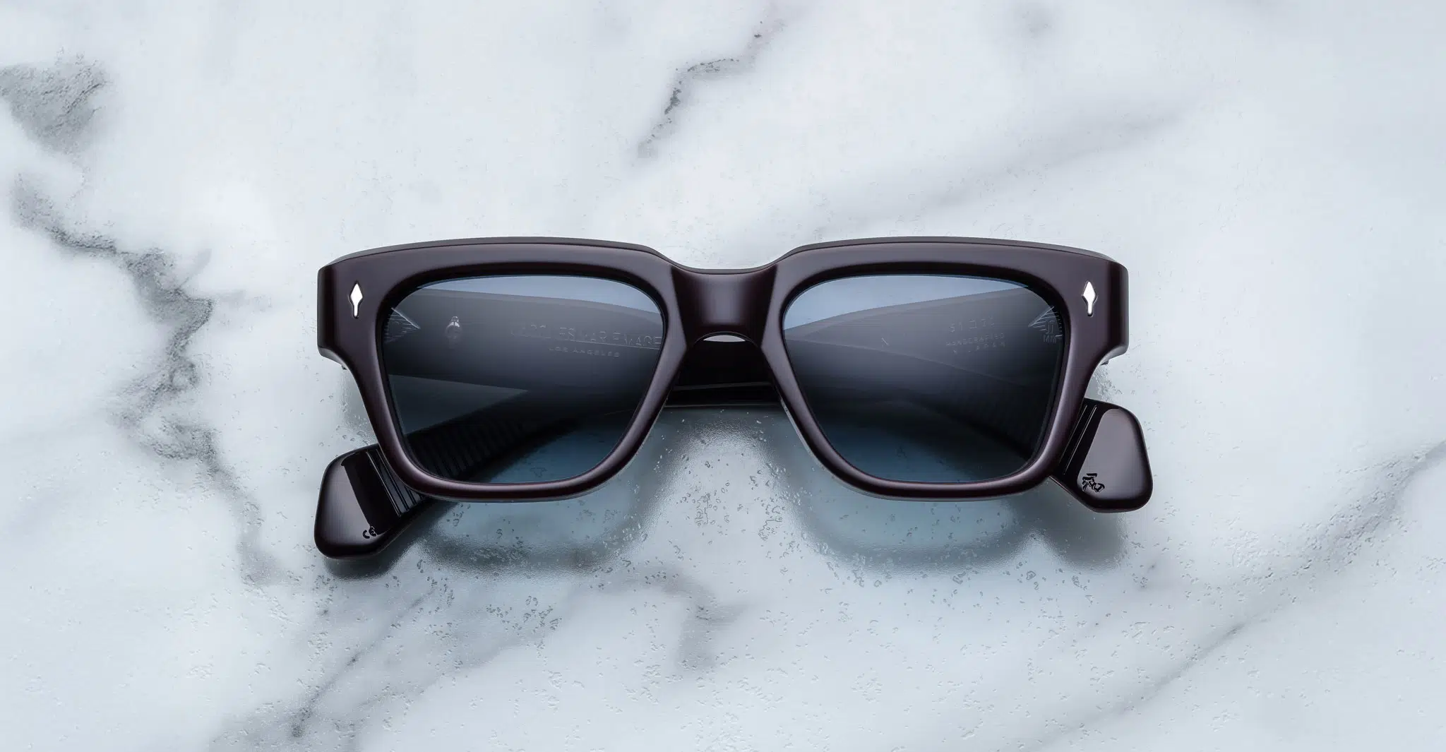 A pair of Jacques Marie Mage Fellini black rectangular sunglasses with dark lenses is laid flat on a white marble surface with gray veining.
