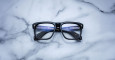 The Jacques Marie Mage Yves black rectangular eyeglasses with blue-tinted lenses rest on a white marble surface accented with gray veining.