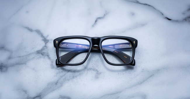 The Jacques Marie Mage Yves black rectangular eyeglasses with blue-tinted lenses rest on a white marble surface accented with gray veining.