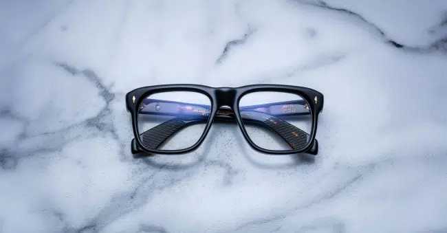 The Jacques Marie Mage Yves black rectangular eyeglasses with blue-tinted lenses rest on a white marble surface accented with gray veining.
