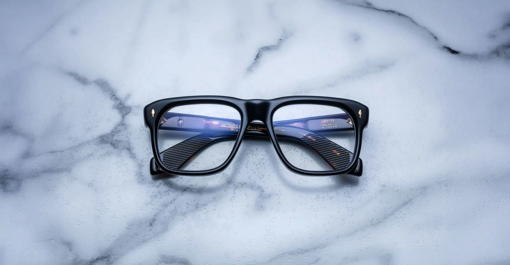The Jacques Marie Mage Yves black rectangular eyeglasses with blue-tinted lenses rest on a white marble surface accented with gray veining.