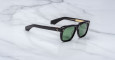 The Jacques Marie Mage Yves sunglasses, featuring a black rectangular frame, green tinted lenses, and gold detailing near the hinges, are displayed on a white marble surface.