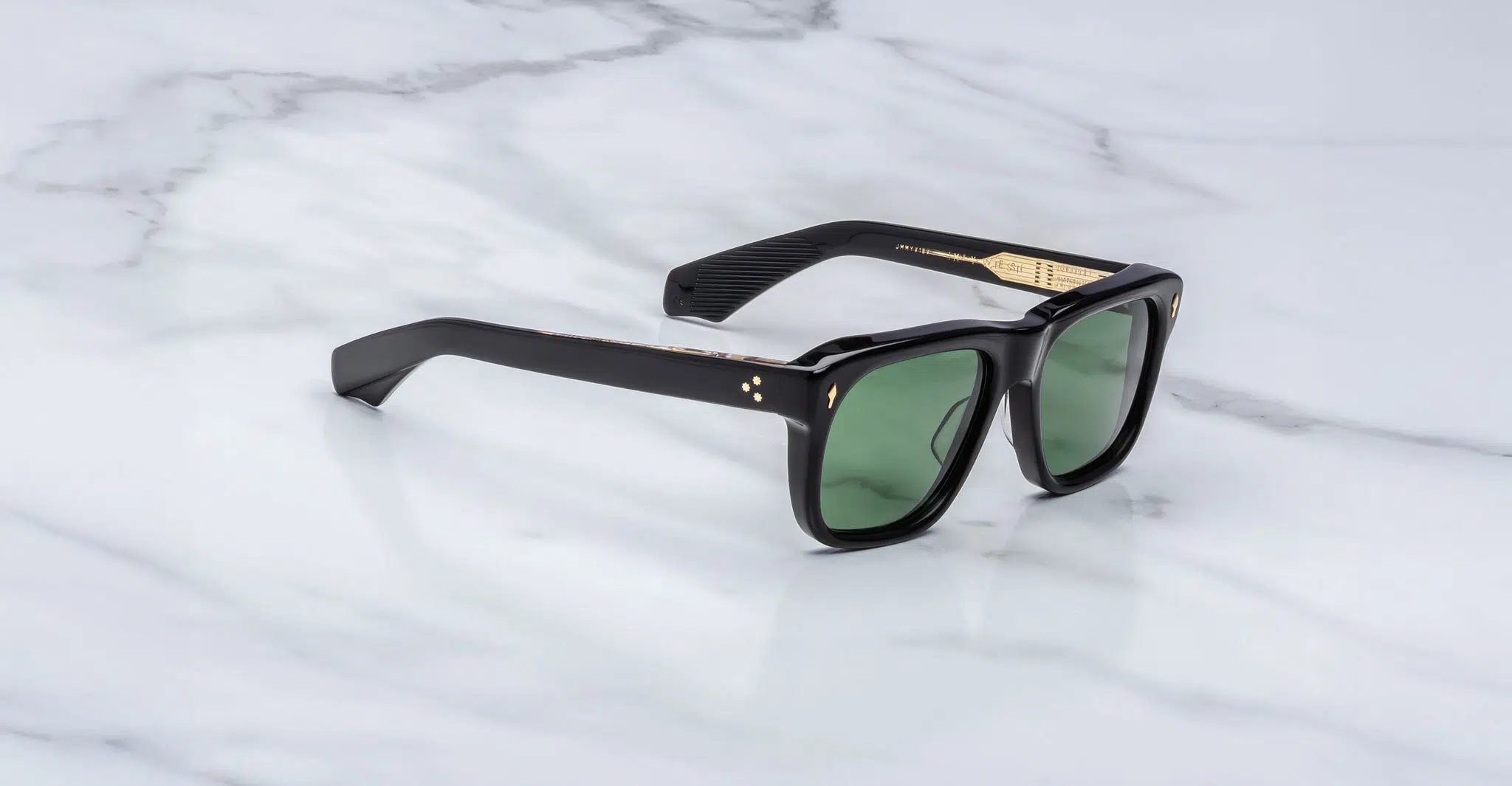 The Jacques Marie Mage Yves sunglasses, featuring a black rectangular frame, green tinted lenses, and gold detailing near the hinges, are displayed on a white marble surface.