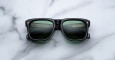 A pair of Jacques Marie Mage Yves black rectangular sunglasses with dark lenses rests on a white marble surface, their lenses reflecting part of a building with horizontal lines.