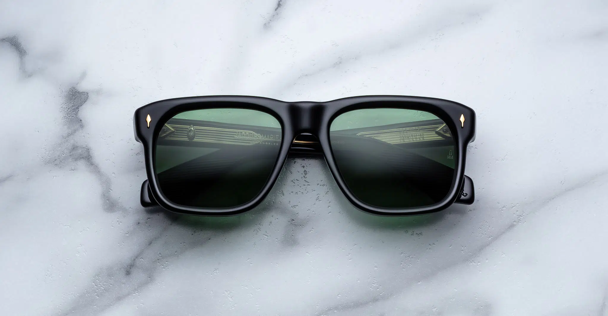 A pair of Jacques Marie Mage Yves black rectangular sunglasses with dark lenses rests on a white marble surface, their lenses reflecting part of a building with horizontal lines.