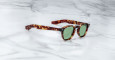 The Jacques Marie Mage Zephirin 47 sunglasses in brown tortoiseshell with green lenses sit on a white marble surface, angled with open temples as light reflects off the smooth background.
