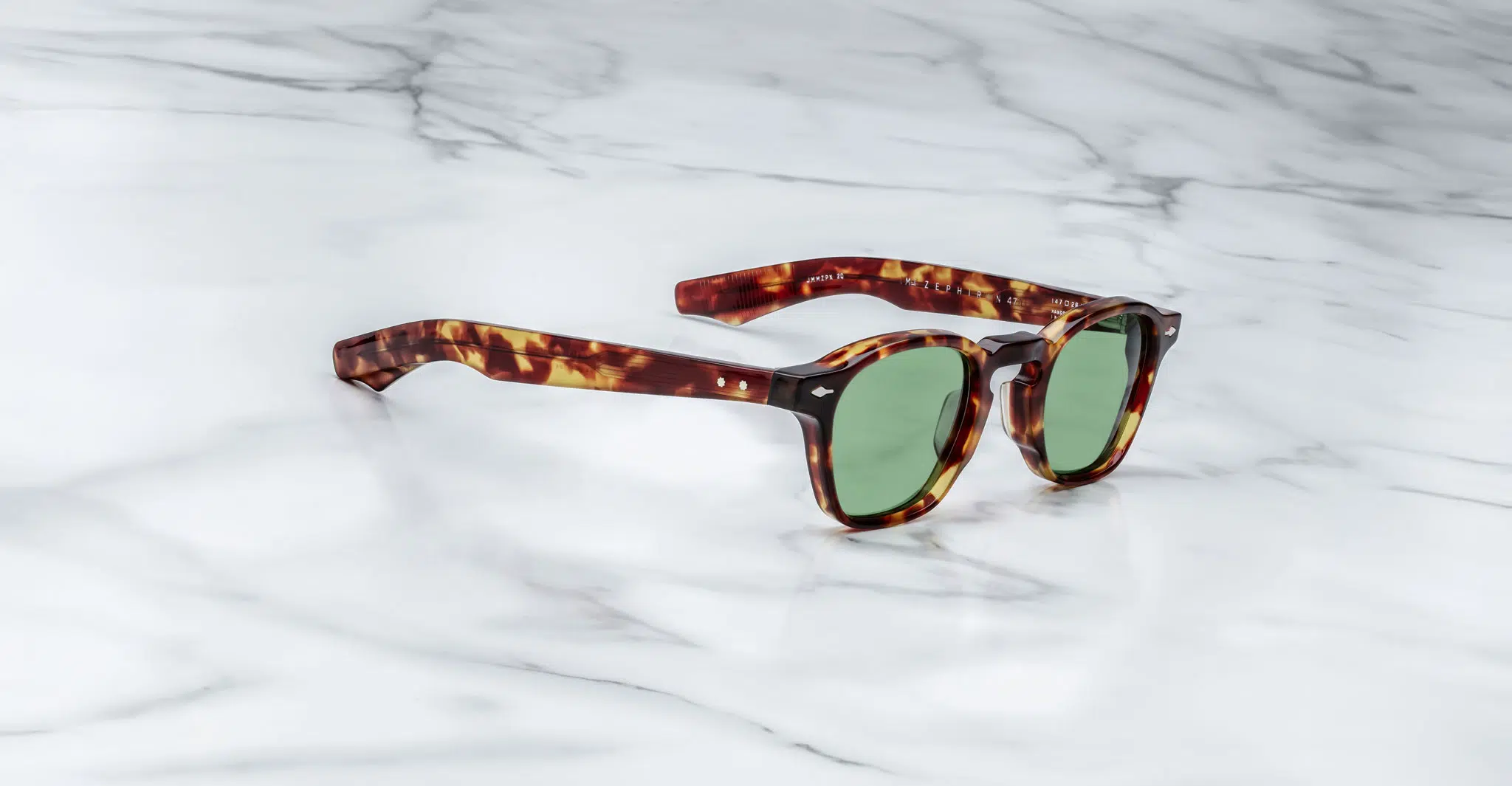 The Jacques Marie Mage Zephirin 47 sunglasses in brown tortoiseshell with green lenses sit on a white marble surface, angled with open temples as light reflects off the smooth background.