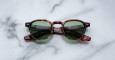 The Jacques Marie Mage Zephirin 47 tortoiseshell sunglasses with green lenses rest on a white marble surface, their arms folded to reveal subtle brand details inside.