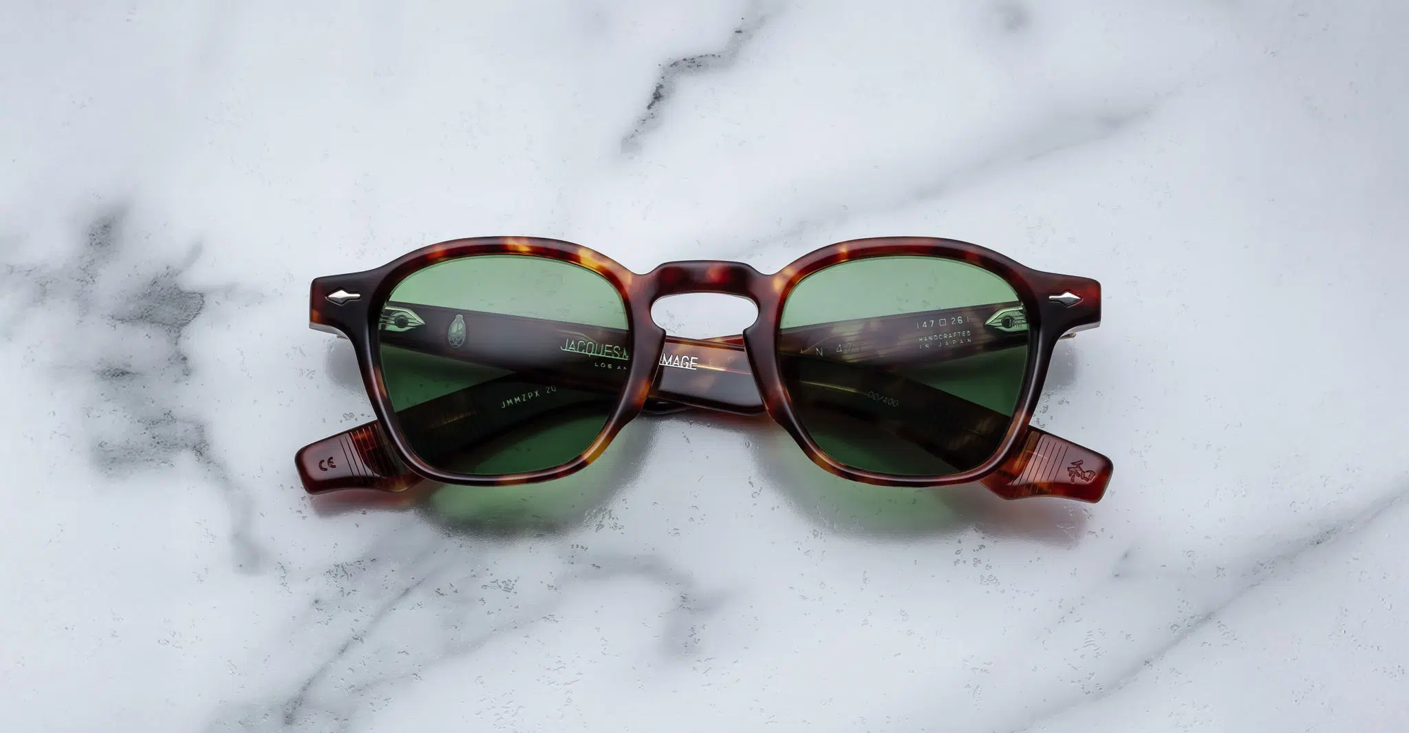 The Jacques Marie Mage Zephirin 47 tortoiseshell sunglasses with green lenses rest on a white marble surface, their arms folded to reveal subtle brand details inside.