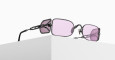 A pair of black metal sunglasses with rectangular, light purple-tinted lenses rest on the edge of a white surface, with unique side shields attached to the arms.
