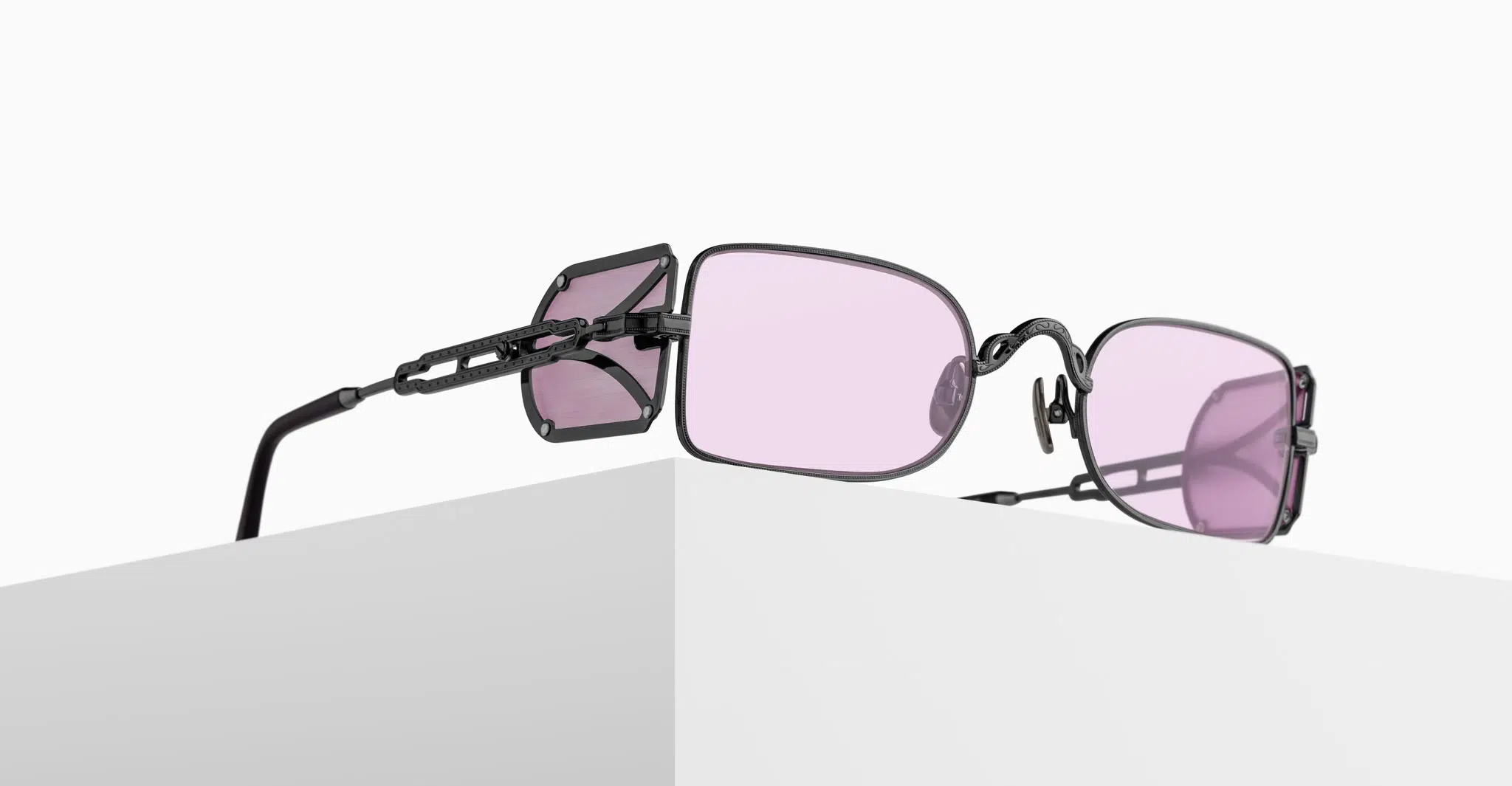 Matsuda-10611H-Ruthenium-10611H.SG.RTM.48-Pedestal-Dan-Deutsch A pair of black metal sunglasses with rectangular, light purple-tinted lenses rest on the edge of a white surface, with unique side shields attached to the arms.