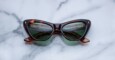 A pair of cat-eye sunglasses with dark green lenses and a brown tortoiseshell frame rests on a light gray marble surface.