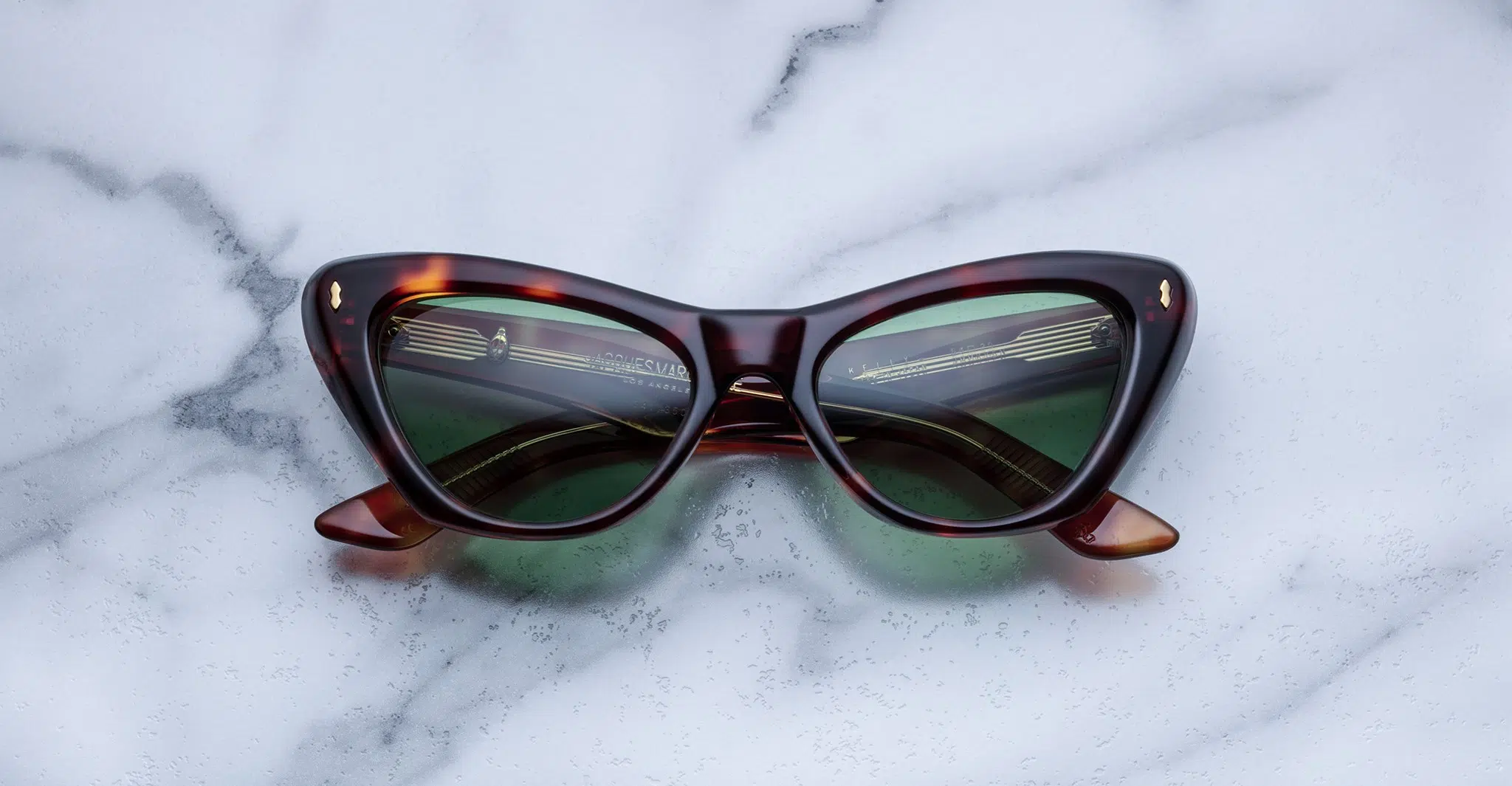 A pair of cat-eye sunglasses with dark green lenses and a brown tortoiseshell frame rests on a light gray marble surface.
