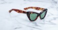 A pair of tortoiseshell cat-eye sunglasses with green lenses is placed on a white marble surface.