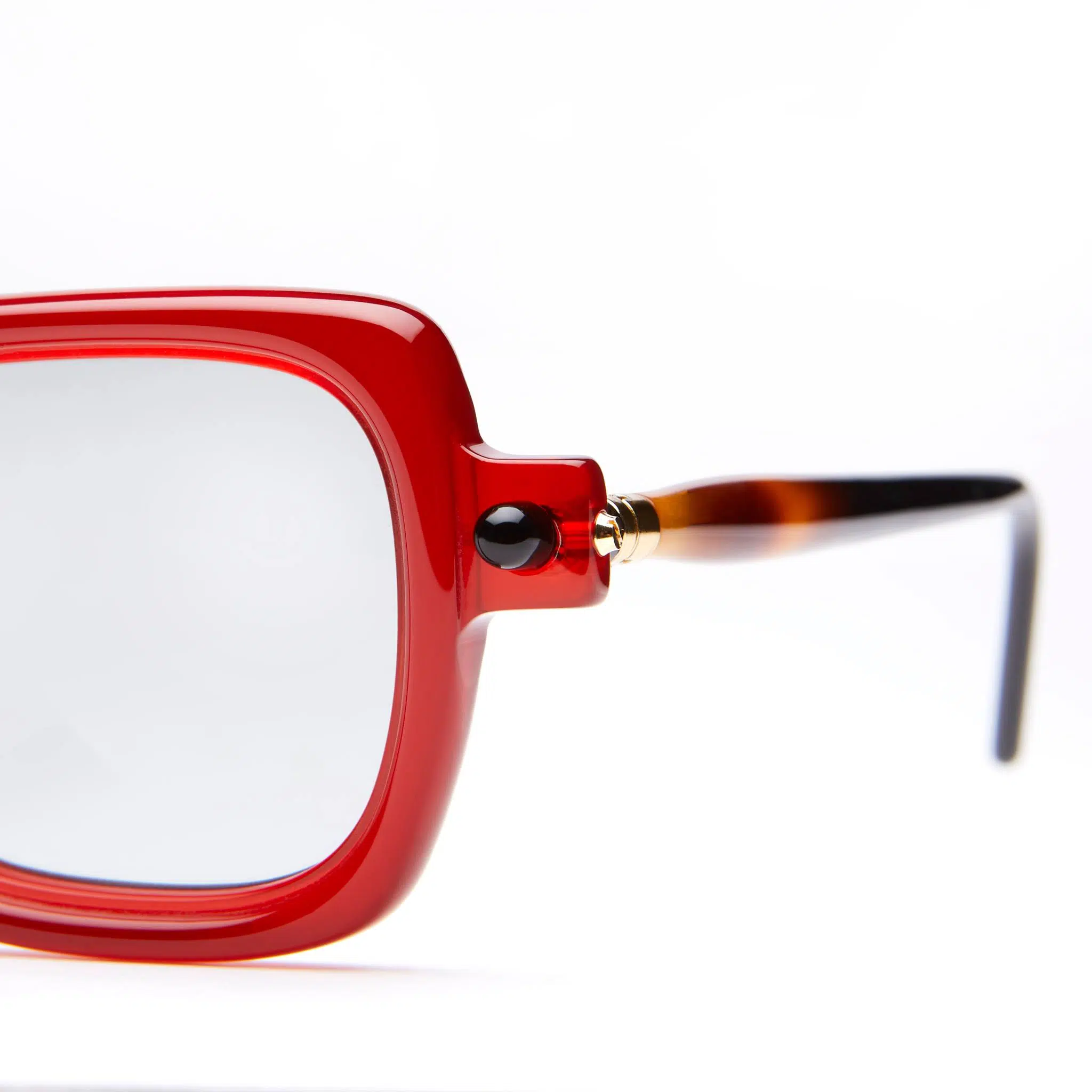 Close-up of the left side of a pair of eyeglasses with a thick, glossy red frame, clear lens, black hinge detail, and tortoiseshell-patterned temple against a white background.
