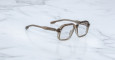 Jacques Marie Mage Domoto transparent gray square-framed eyeglasses with thick rims rest on a white marble surface.