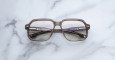 A pair of Jacques Marie Mage Domoto square, translucent brown eyeglasses with thick frames rests on a white marble surface with subtle grey veining; their temples are crossed over each other.