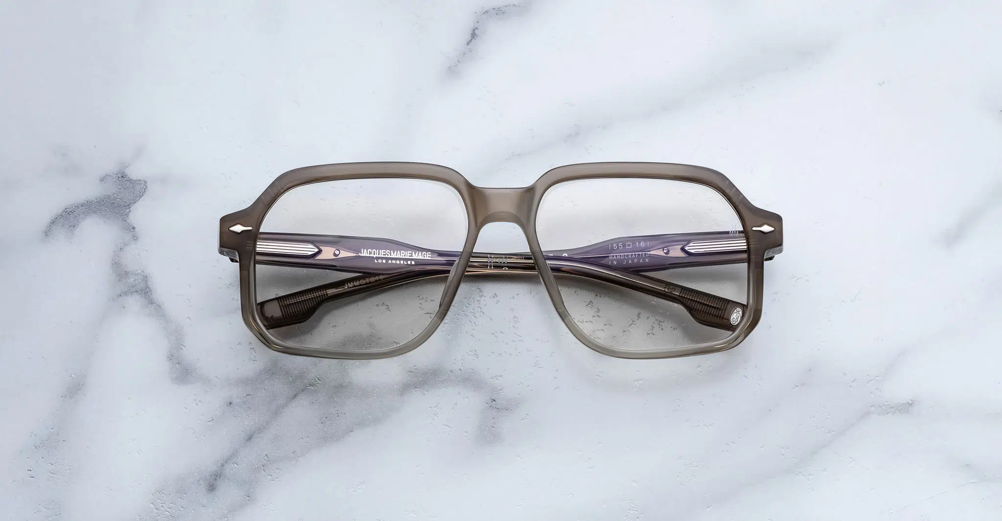 A pair of Jacques Marie Mage Domoto square, translucent brown eyeglasses with thick frames rests on a white marble surface with subtle grey veining; their temples are crossed over each other.