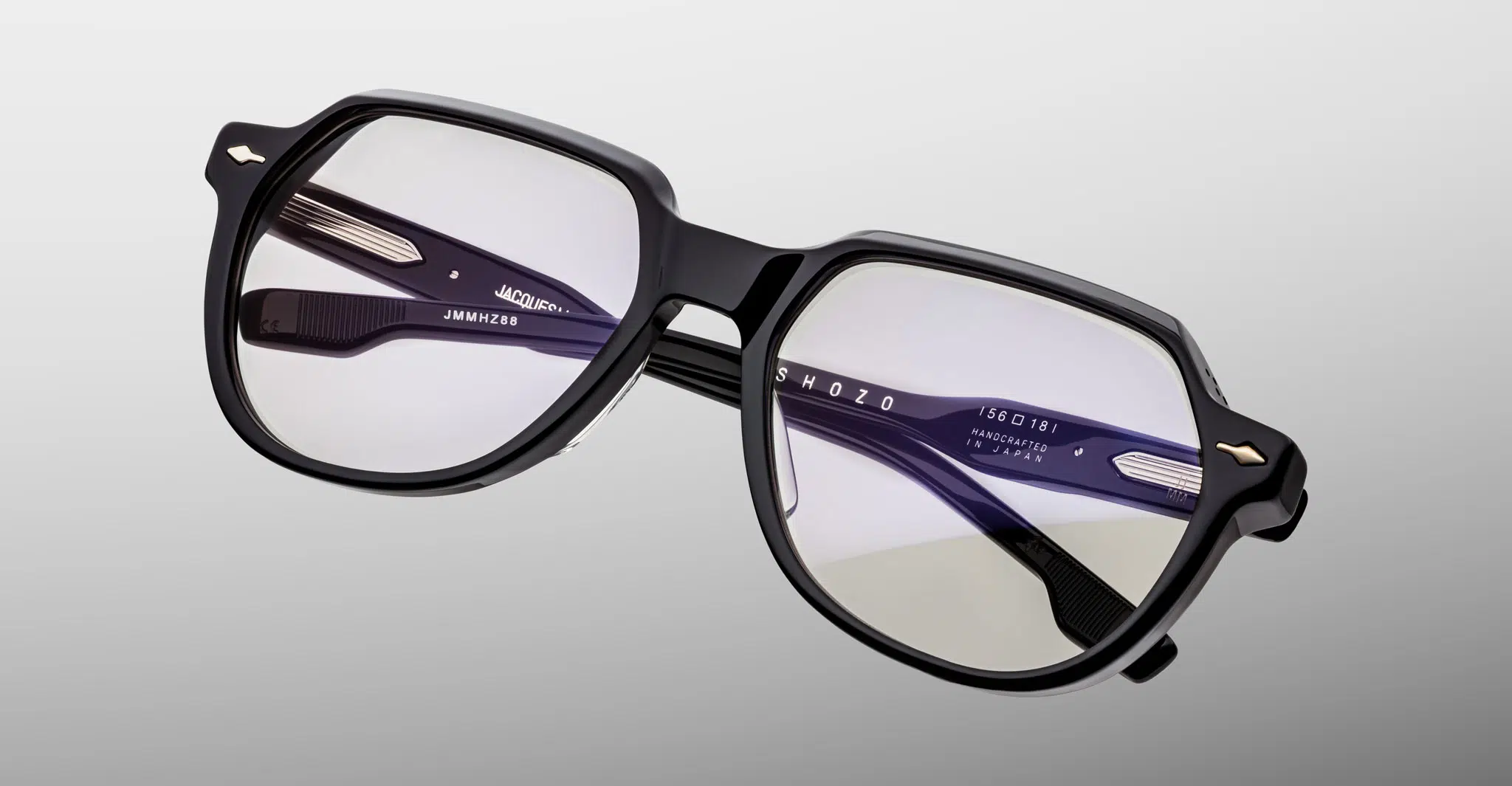 Jacques-Marie-Mage-Shozo-Black-RX-JMMHZRX-88-Detail1-Dan-Deutsch The Jacques Marie Mage Shozo features black-rimmed eyeglasses with clear lenses and a modern geometric frame, displayed against a light gray gradient background. Branding text appears on the inside of the temples.