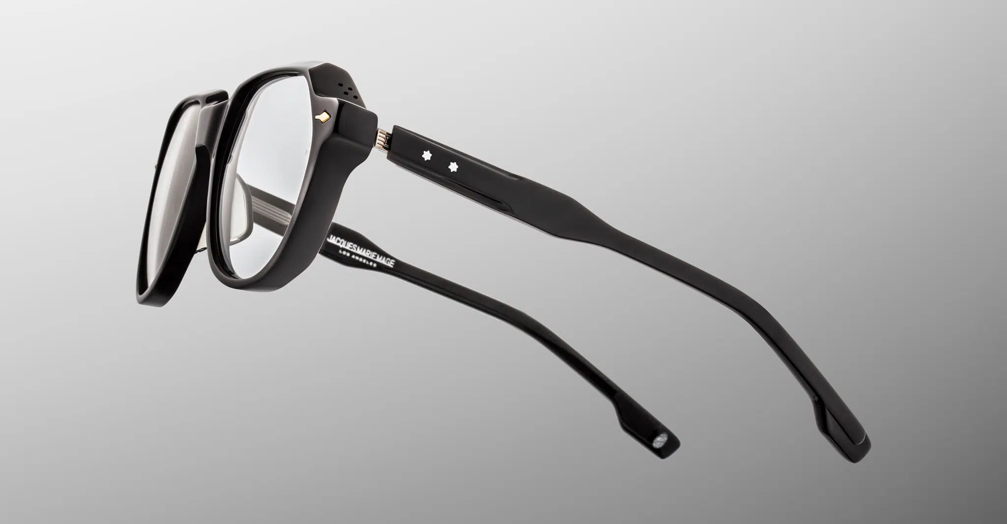 Jacques-Marie-Mage-Shozo-Black-RX-JMMHZRX-88-Detail2-Dan-Deutsch The Jacques Marie Mage Shozo eyeglasses feature black thick-rimmed frames with clear lenses, viewed from an angled side perspective on a light gray background. Two small white star accents adorn the temples.