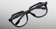 The Jacques Marie Mage Shozo glasses, black with thick rims and folded arms, feature small white star-shaped accents on the arms and are shown from above against a light gray gradient background.