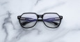 The Jacques Marie Mage Shozo eyeglasses, featuring black frames and clear lenses, rest on a white marble surface. The temples display brand and size details, and the frames have a subtle shine.