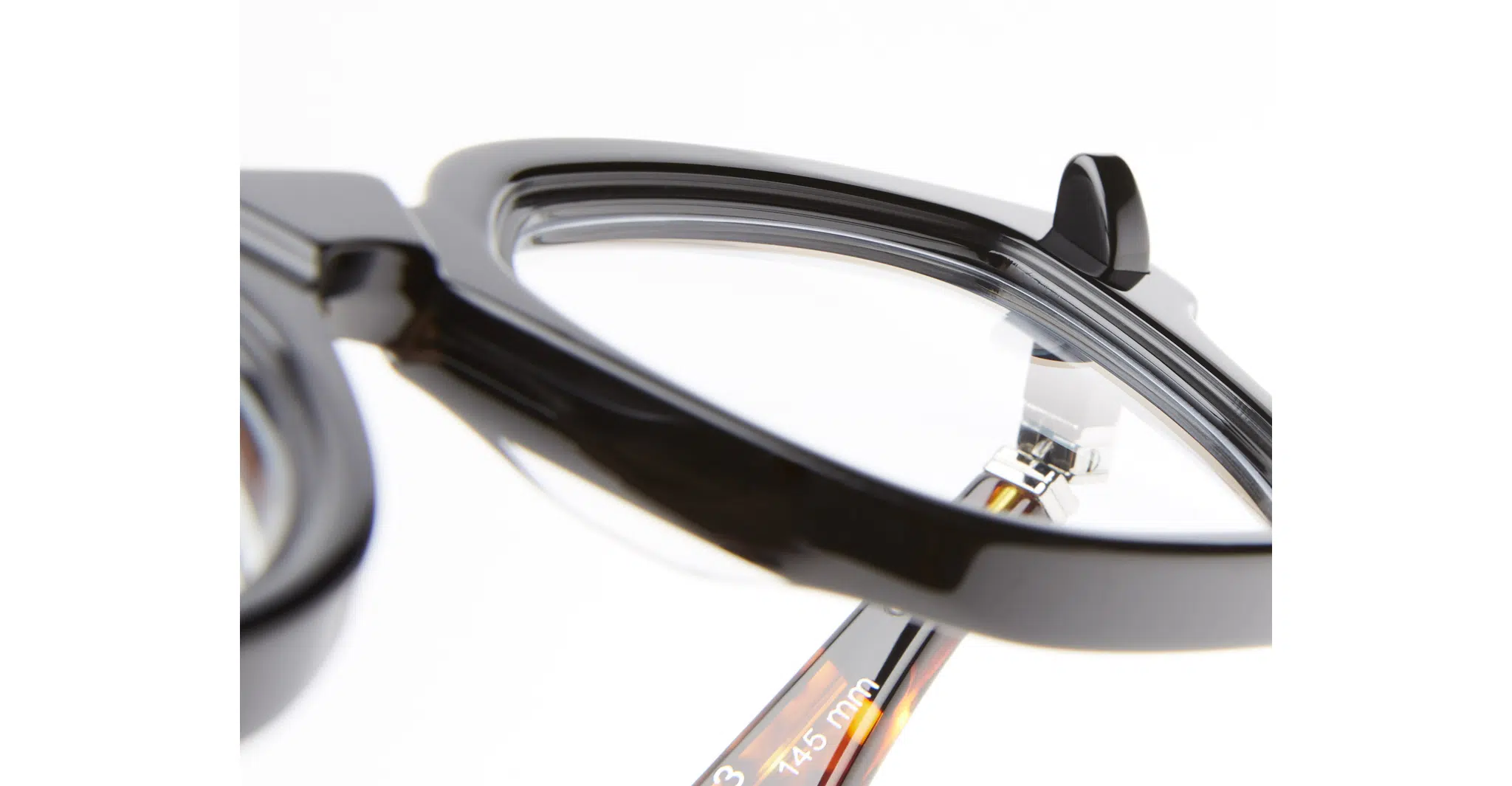 Kuboraum-Q3-BST-Black-Shine-KRO0Q3BST00000OP-Detail1-Dan-Deutsch Close-up of the Kuboraum Q3 black eyeglasses’ corner, highlighting the glossy frame, clear lens, and part of the temple arm with measurement details visible against a white background.