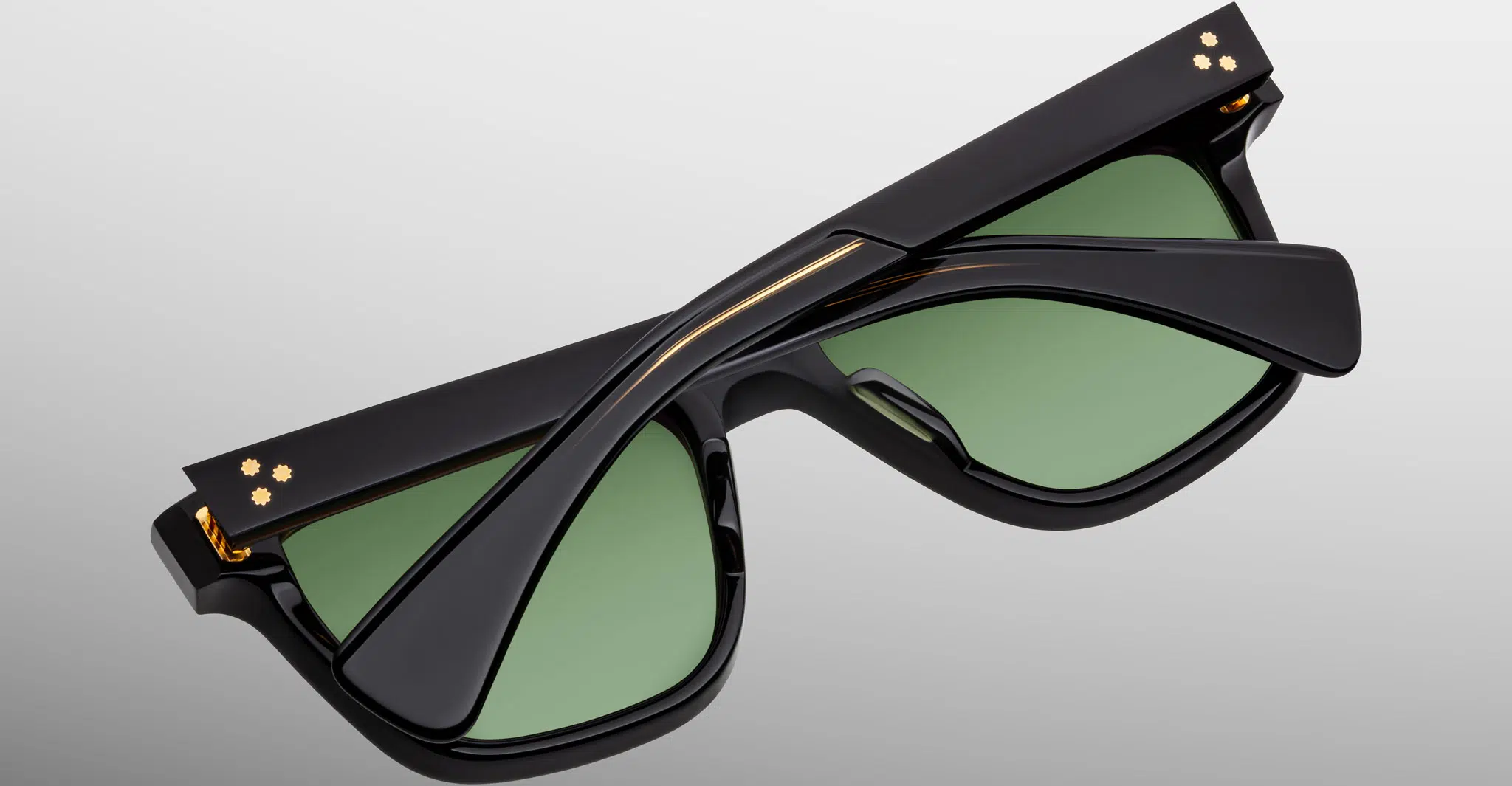 The Jacques Marie Mage Lankaster sunglasses are shown folded neatly, featuring black frames with green lenses and gold accents near the hinges, set against a plain light gray background.